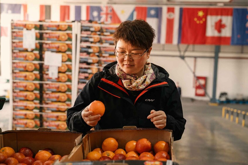 A person holding an orange, standing in front of a box filled with oranges, in a store with various flags in the background. 4