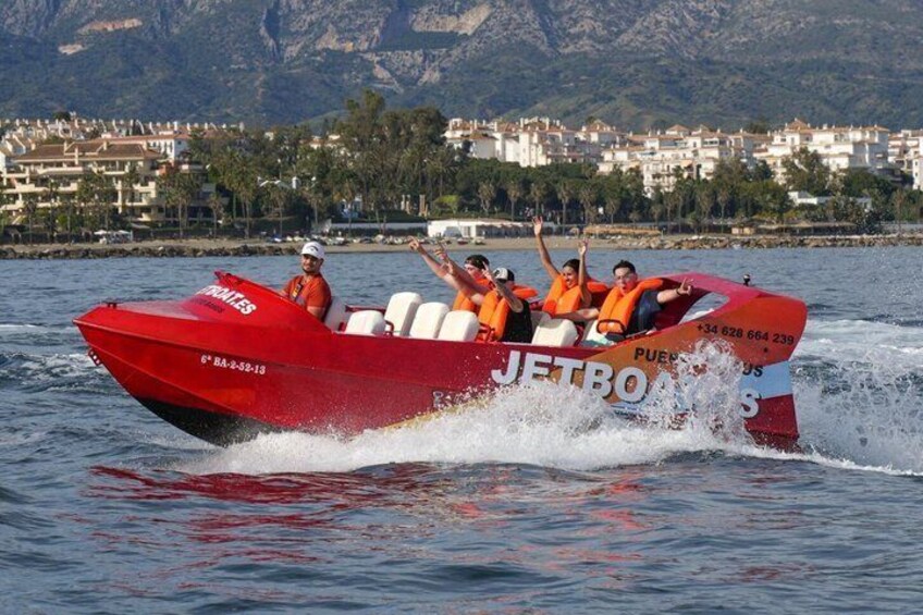 A red speedboat with passengers wearing orange life jackets, moving swiftly on water with a mountainous coastline in the background. 3