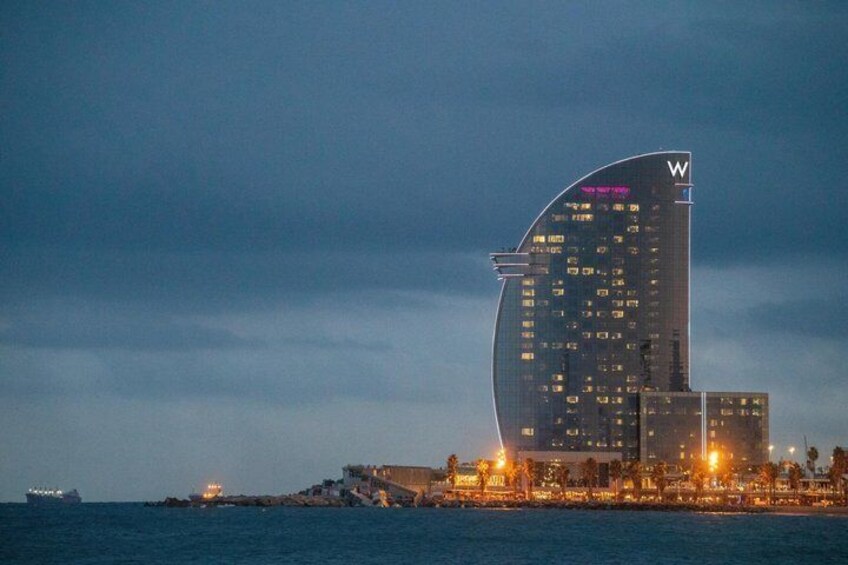 A curved skyscraper with illuminated windows, a waterfront, and a ship in the background. 1