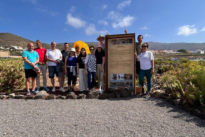 A group of people standing in front of a sign with text and images, with a clear sky and distant hills in the background. 5