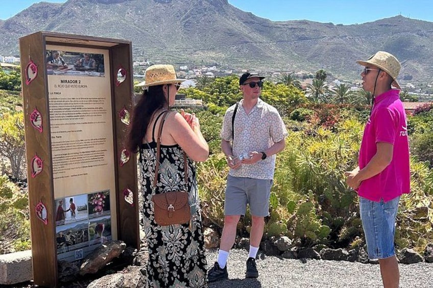 A group of people standing near a signboard with text and images, with a mountainous landscape in the background. 4