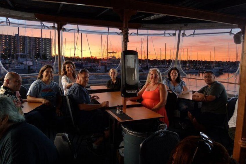 A group of people seated at tables on a boat with a marina and sailboats in the background. 1