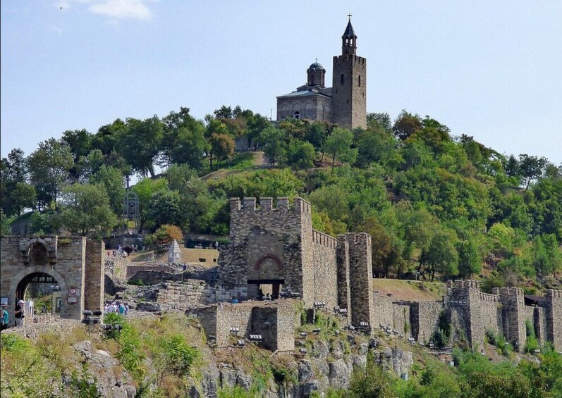 A historic hilltop fortress with a church and bell tower, surrounded by trees and ruins. 2