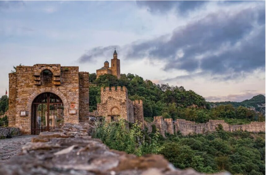 An ancient stone fortress with a gate, surrounded by lush greenery and a cloudy sky. 1
