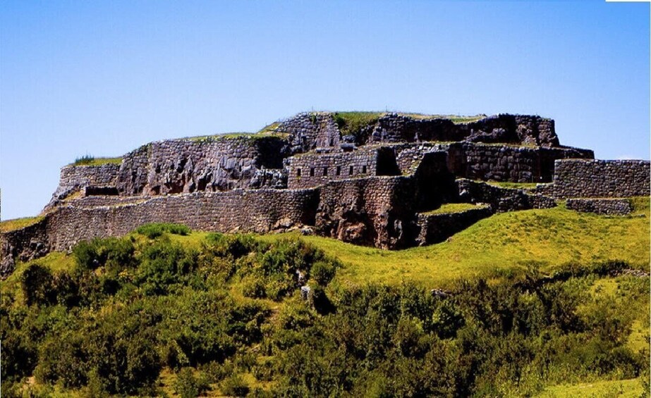 Una gran estructura de piedra antigua sobre una colina, con vegetación verde y densa en primer plano. 5