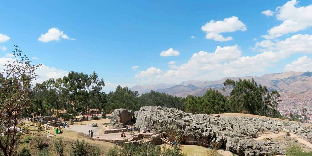 Un sitio arqueológico rocoso con visitantes, rodeado de árboles y montañas al fondo. 3