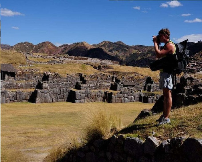 Una persona fotografiando antiguas ruinas de piedra con montañas al fondo. 1