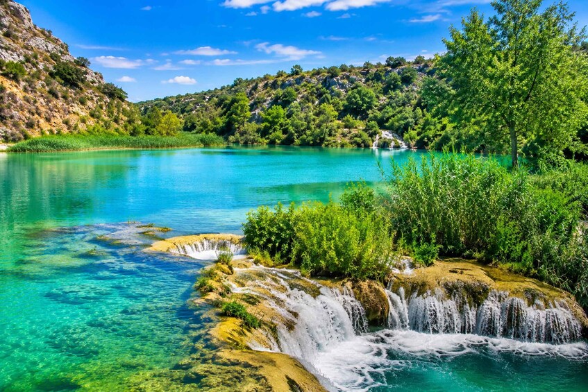 A turquoise lake with a waterfall, surrounded by lush greenery and rocky hills. 1