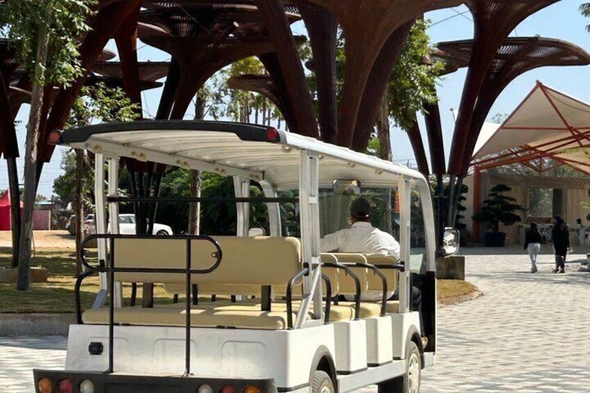 A white golf cart with a beige seat, parked under a large tree with a metal canopy structure in the background. 3