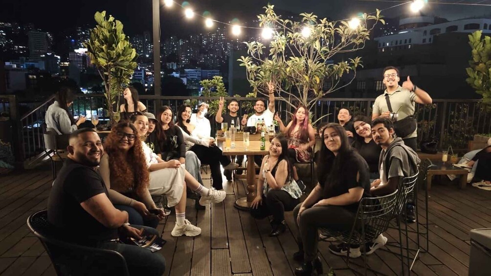 A group of people gathered around a table on a wooden deck at night, with city lights in the background. 1