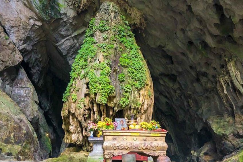 A cave with a rock formation covered in green moss, a small altar with offerings, and a carved stone base. 2