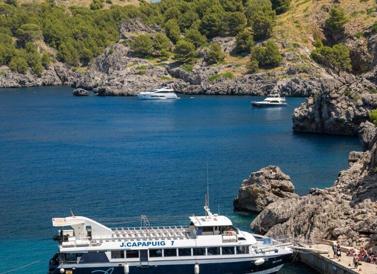 A ferry named 'J. Capapuig 7' docked at a rocky harbor with clear blue water and a mountainous backdrop. 2
