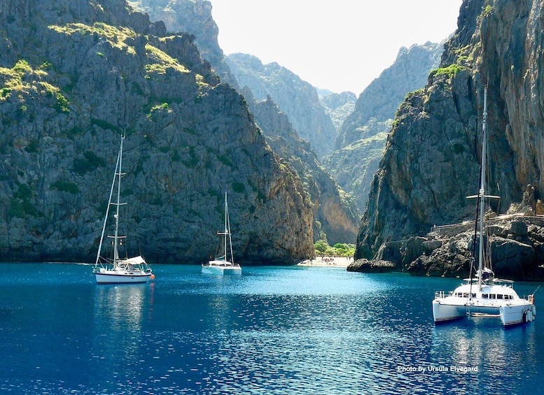 A coastal scene with three sailboats anchored in calm waters surrounded by rocky cliffs. 1