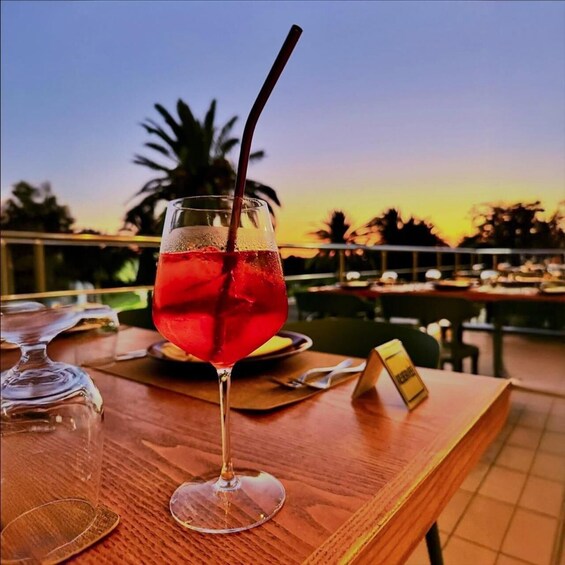 A glass of red cocktail with a straw on a wooden table with a sunset in the background. 2