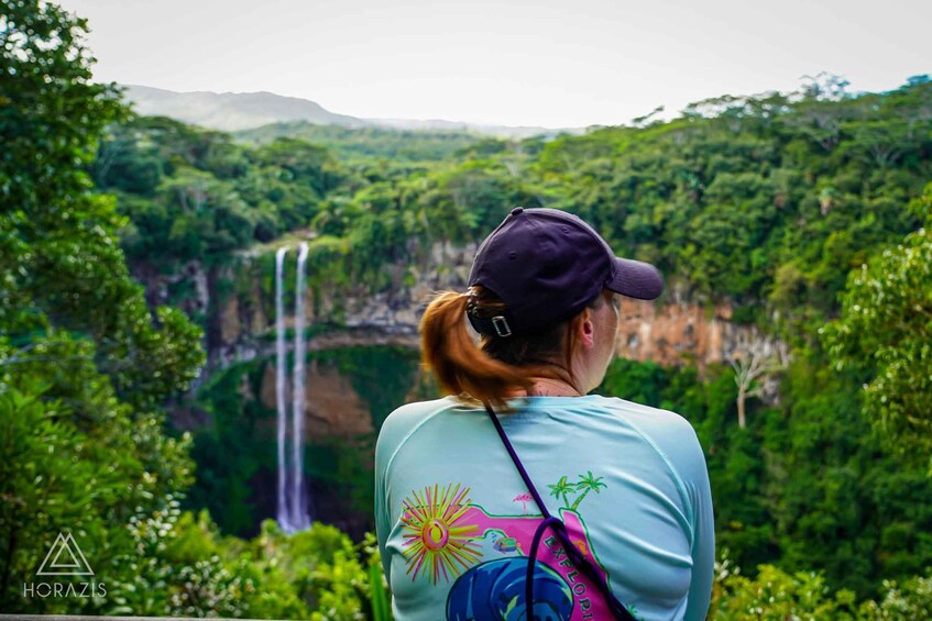 A person wearing a cap and a light blue shirt with a colorful design, overlooking a lush green landscape with a waterfall. 1