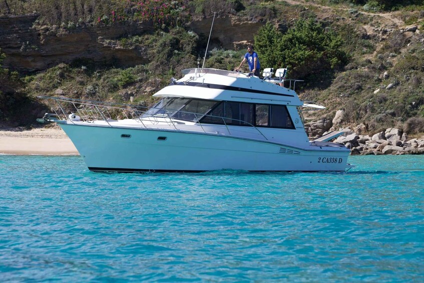 A white yacht with the registration number 2 CA338 D on turquoise water, with a rocky coastline and sparse vegetation in the background. 3