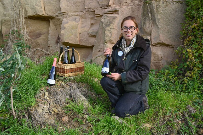 A person kneeling on grass holding a wine bottle, with several bottles and a basket of wine bottles in the background. 2