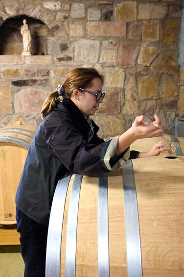 A person pouring a liquid from a glass into a wooden barrel. 5