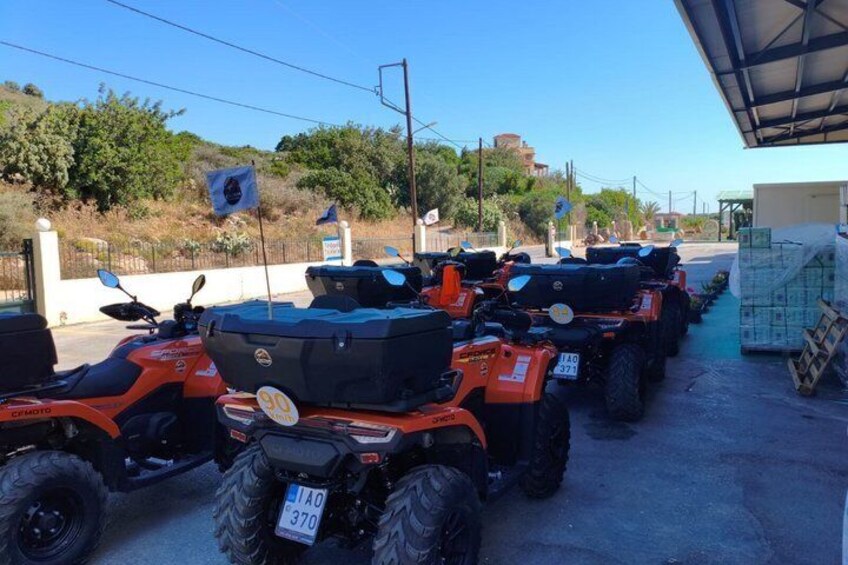 A row of orange ATVs parked in a lot with a clear sky and greenery in the background. 4