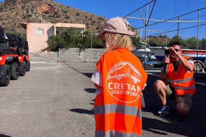 Two people in orange vests with 'CRETAN QUADVENTURES' logo, a red ATV, and a boat in the background. 5