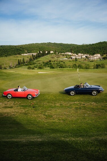 Two vintage cars parked on a golf course with a green hill and buildings in the background. 2
