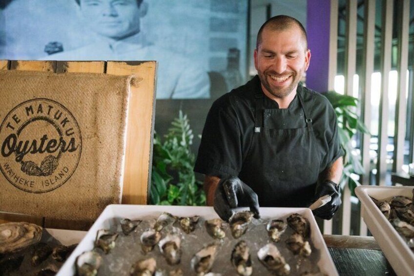 A person handling oysters in a kitchen setting with a branded box labeled 'TE MATUKU Oysters.' 1