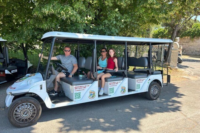 A golf cart with three passengers, parked on a paved area with trees in the background. 1