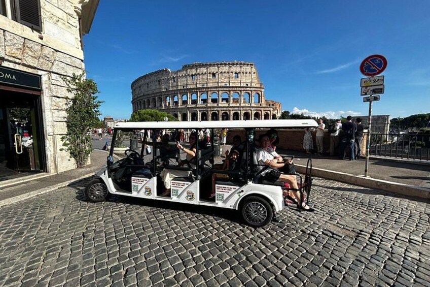 A white golf cart with passengers, a cobblestone street, and the Colosseum in Rome. 3