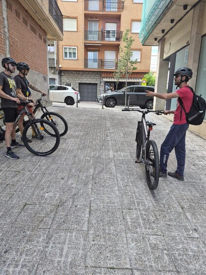 A group of people with bicycles on a city street, a parked car, and a building with balconies. 2