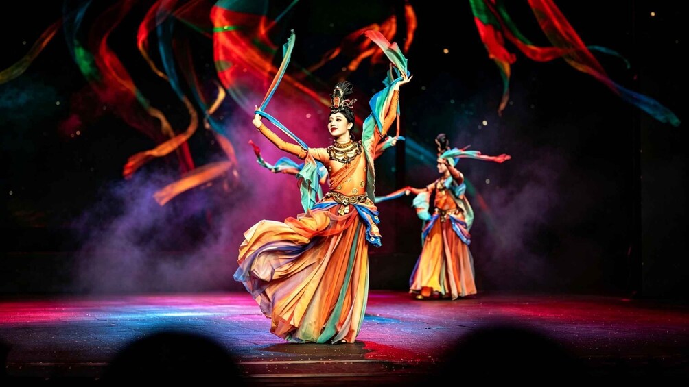 A performer in traditional costume with flowing fabric, on a stage with a dark background and colorful lighting. 2