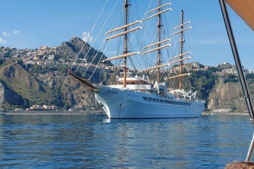 A large sailing ship with multiple masts and sails, docked near a coastal town with hills in the background. 5