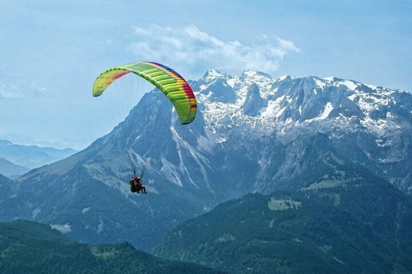 A paraglider with a multicolored parachute flying over a snow-capped mountain range. 1