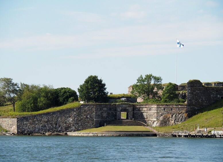 A stone fortification by a body of water, a flagpole with a flag, and lush greenery. 2