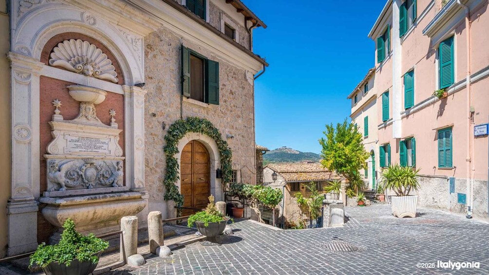A cobblestone street with historic buildings, a stone fountain, and a clear blue sky. 1