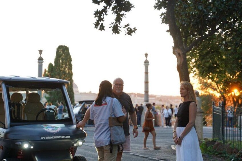 A group of people standing near a vehicle with a website address on the hood, a tree-lined street, and a distant monument. 3