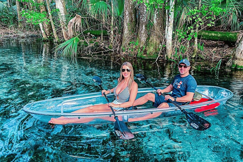 Two people in a transparent kayak on clear water surrounded by trees. 1