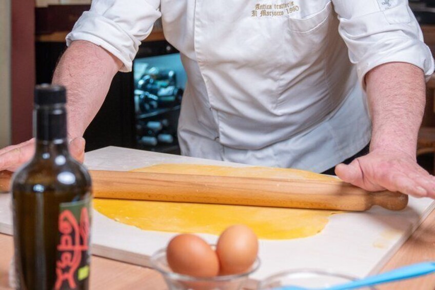 A person rolling dough on a wooden surface with a rolling pin, a bottle of olive oil, and eggs nearby. 4