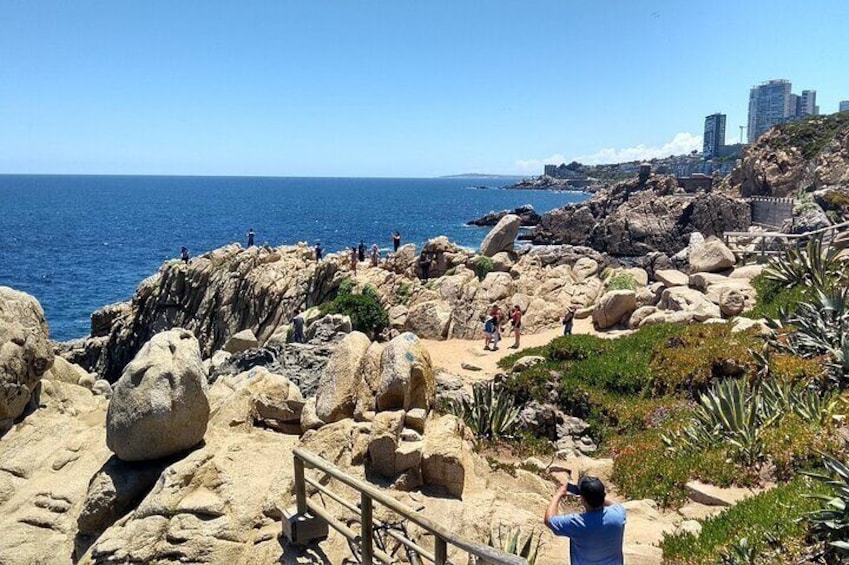 A rocky coastal area with a clear blue sky, a sandy path, and a person taking a photo. 1