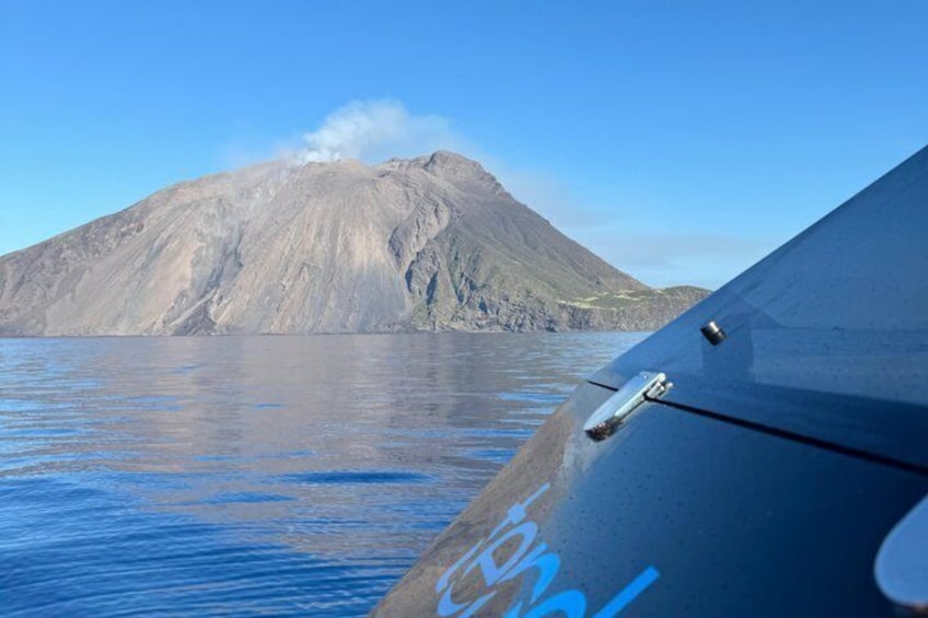 A mountain with a plume of smoke, calm water, and a boat's edge. 1