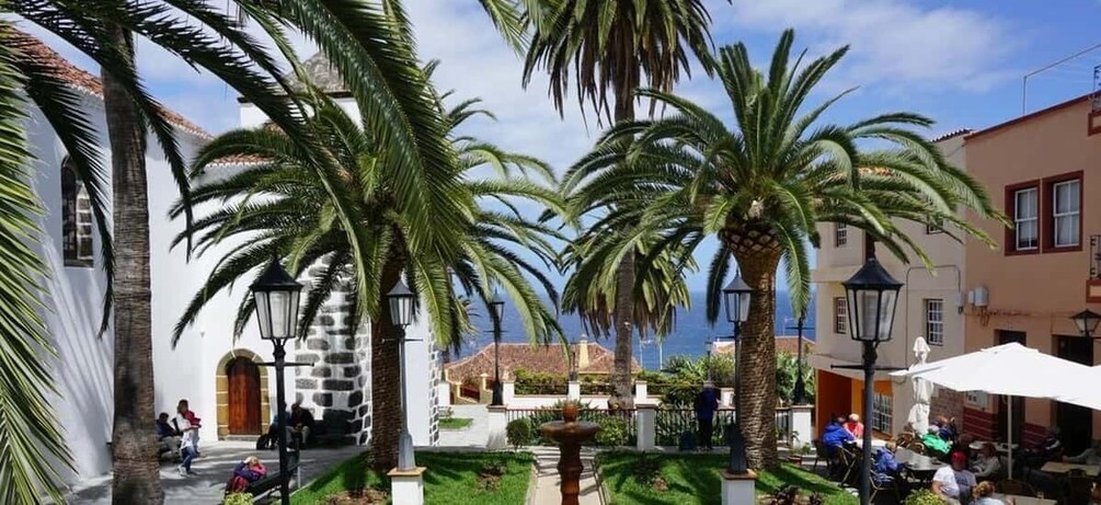 A sunny courtyard with palm trees, a fountain, and people sitting at tables. 1