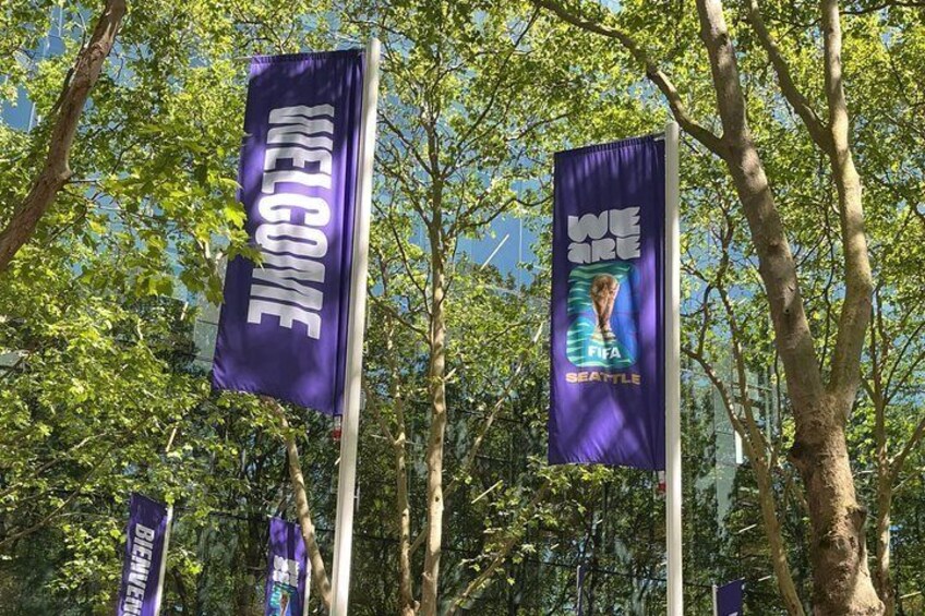 A series of purple banners with 'WELCOME' and 'WE ARE FIFA SEATTLE' text, set against a backdrop of trees. 2
