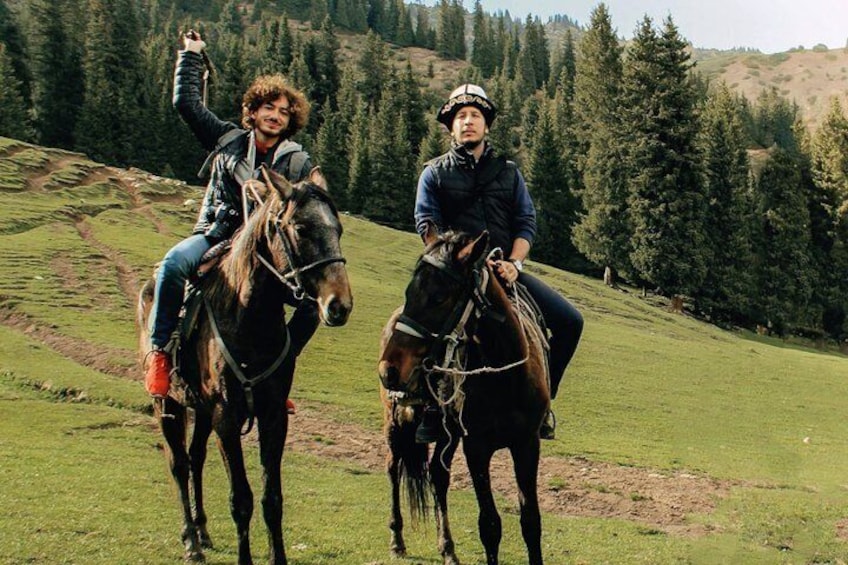 Two people riding horses on a grassy hillside with trees in the background. 1