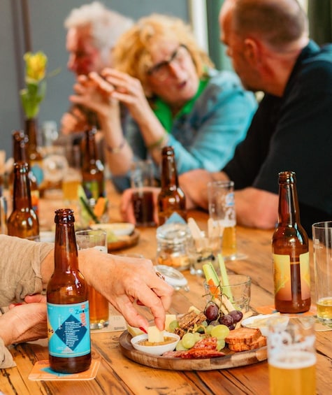 A group of people at a wooden table with beer bottles, glasses, and a variety of food including cheese, grapes, and meats. 3