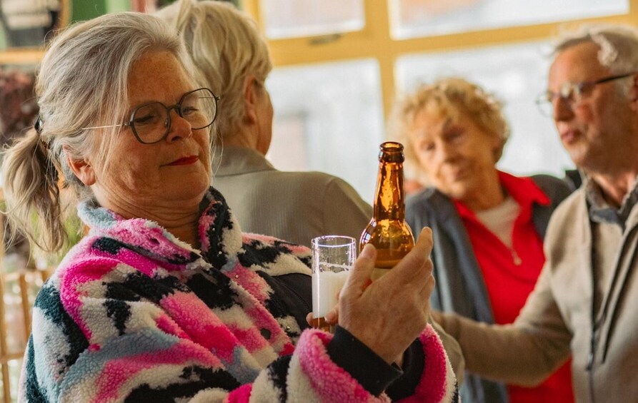 A person holding a bottle and a glass, with other people in the background. 2