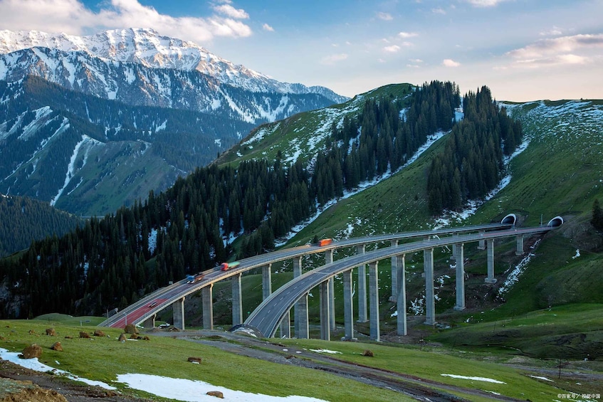 A mountain road with a bridge, snow-capped peaks, and dense forests. 1