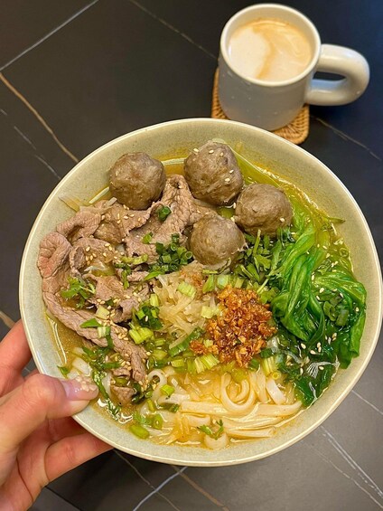A bowl of noodle soup with beef balls, greens, and a cup of coffee. 2