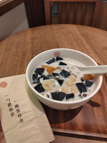 A bowl of dessert with tapioca pearls and fruit pieces, accompanied by a spoon and a napkin with text. 1