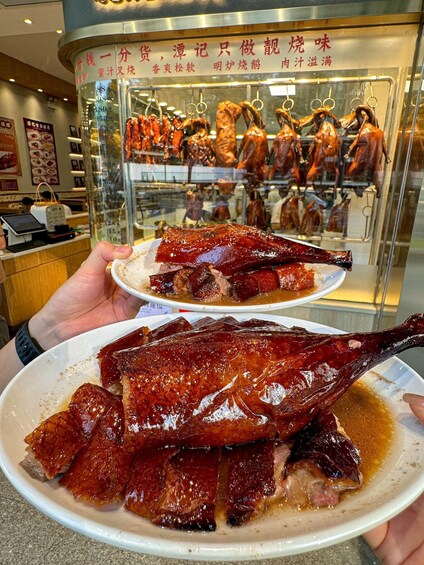 A person holding a plate of roasted duck with crispy skin, in front of a glass display case with more roasted ducks. 5