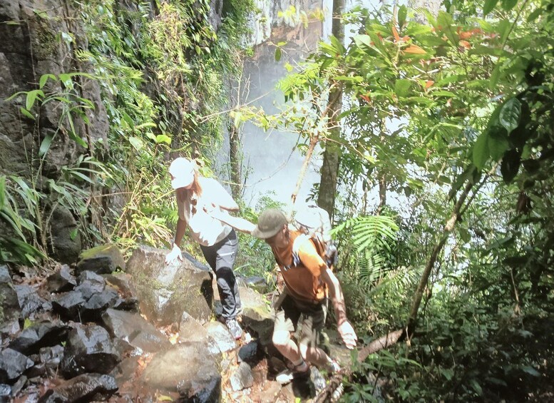 Two hikers navigating a rocky trail in a forested area with dense vegetation and a waterfall in the background. 5