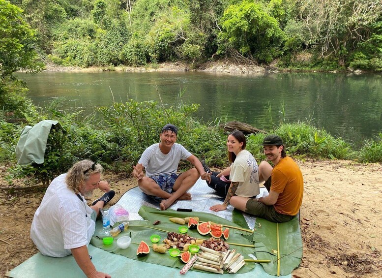 A group of people sitting on a mat by a river, with food and drinks laid out in front of them. 2